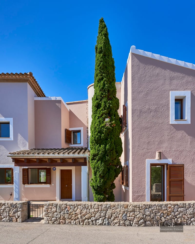 Front view of a pink two-story Mediterranean villa with wooden shutters, a stone wall, and a tall cypress tree.