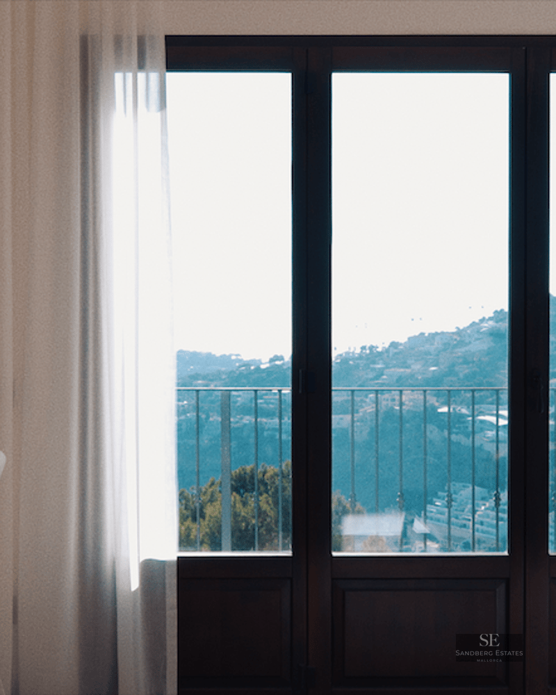 A modern bedroom featuring a desk, a wall-mounted TV, and floor-to-ceiling windows overlooking a scenic hillside and sea.