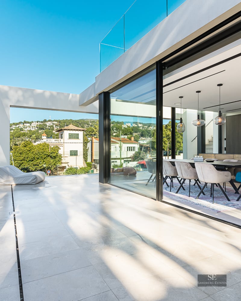 Spacious white terrace with glass railings looking into a modern dining room through large sliding glass doors.