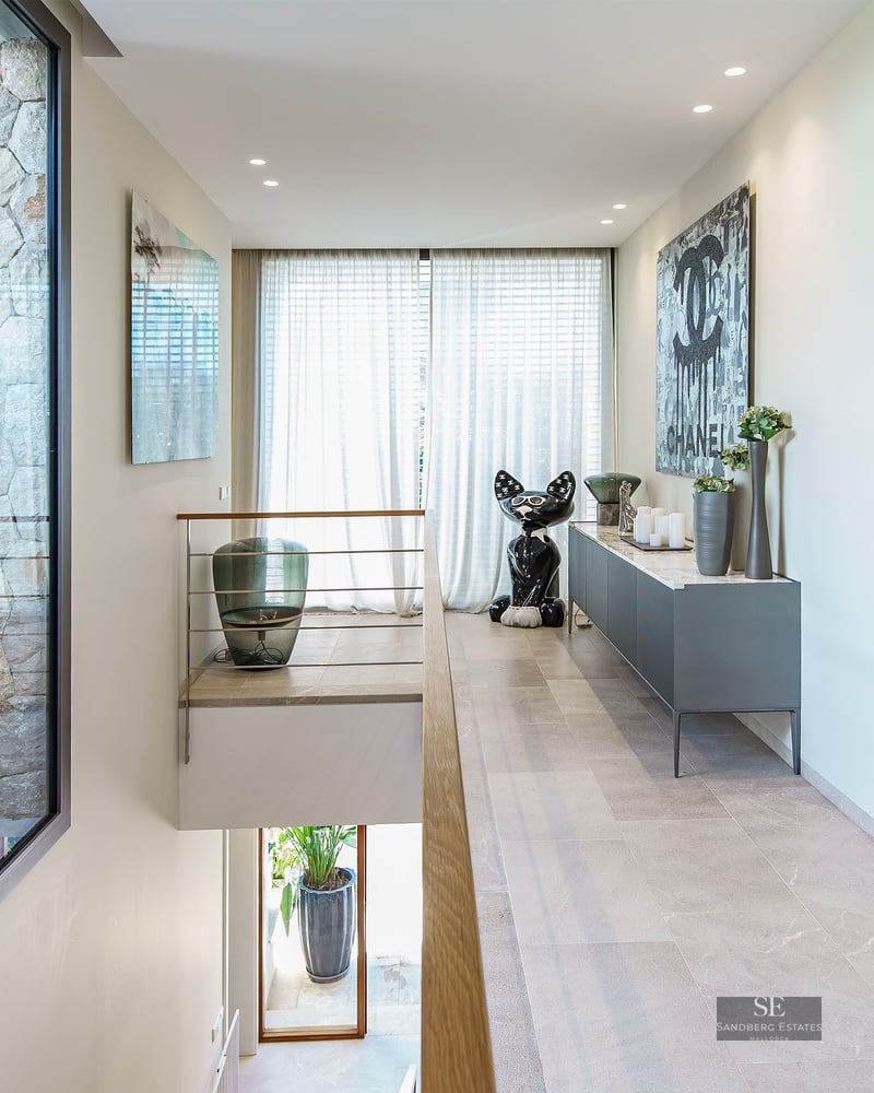 Couloir lumineux, sol en pierre, art moderne et fenêtre donnant sur une cour intérieure avec des plantes.