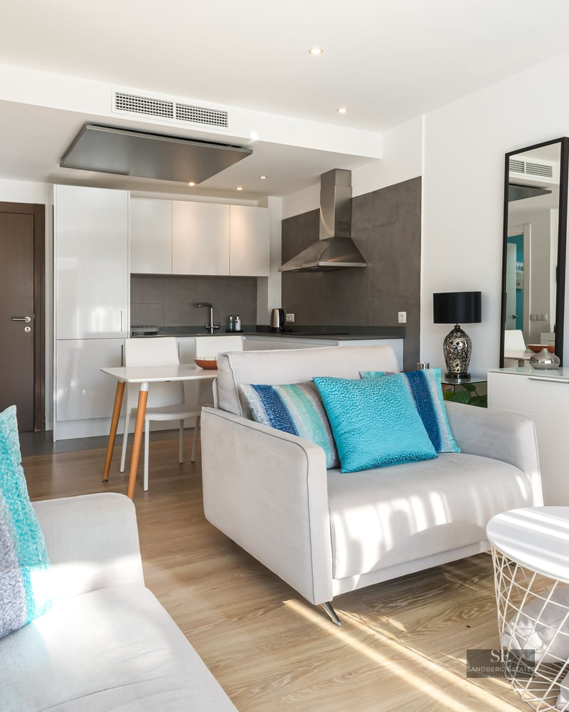 Salon lumineux avec fauteuil gris, coussins turquoise, meuble TV et cuisine moderne en arrière-plan.