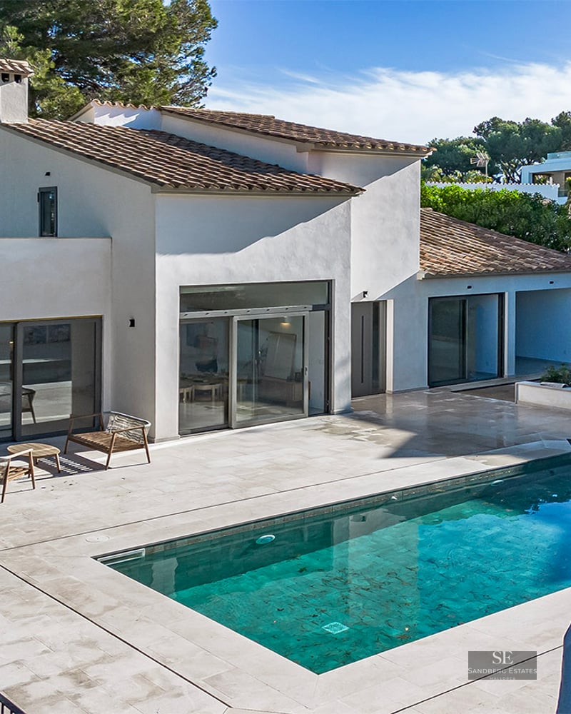 White Mediterranean-style villa with a rectangular blue swimming pool and a large stone deck under bright sunlight.