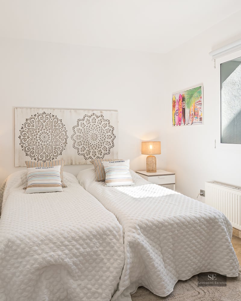 White room with two twin beds, carved wooden headboard, wicker lamps, and a window showing garden views.