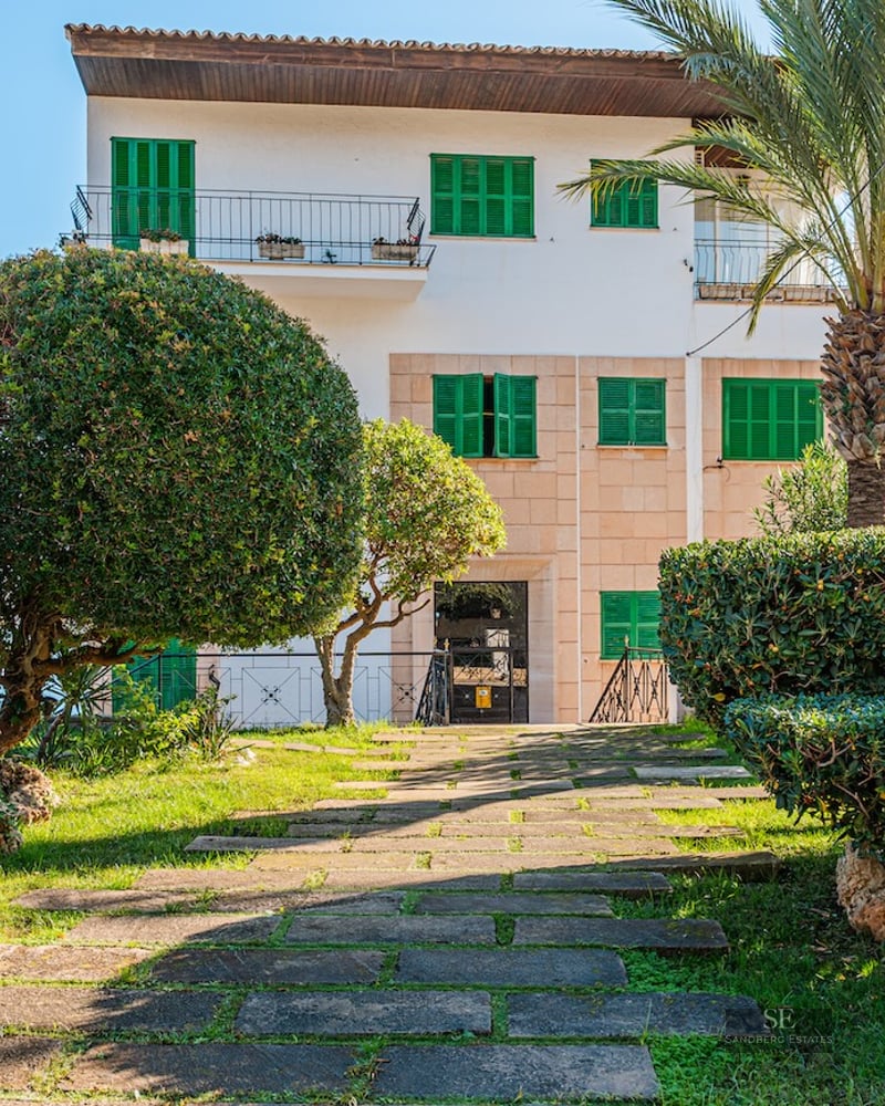 Stone pathway leads through a lush garden with palm trees to a white villa with vibrant green shutters.