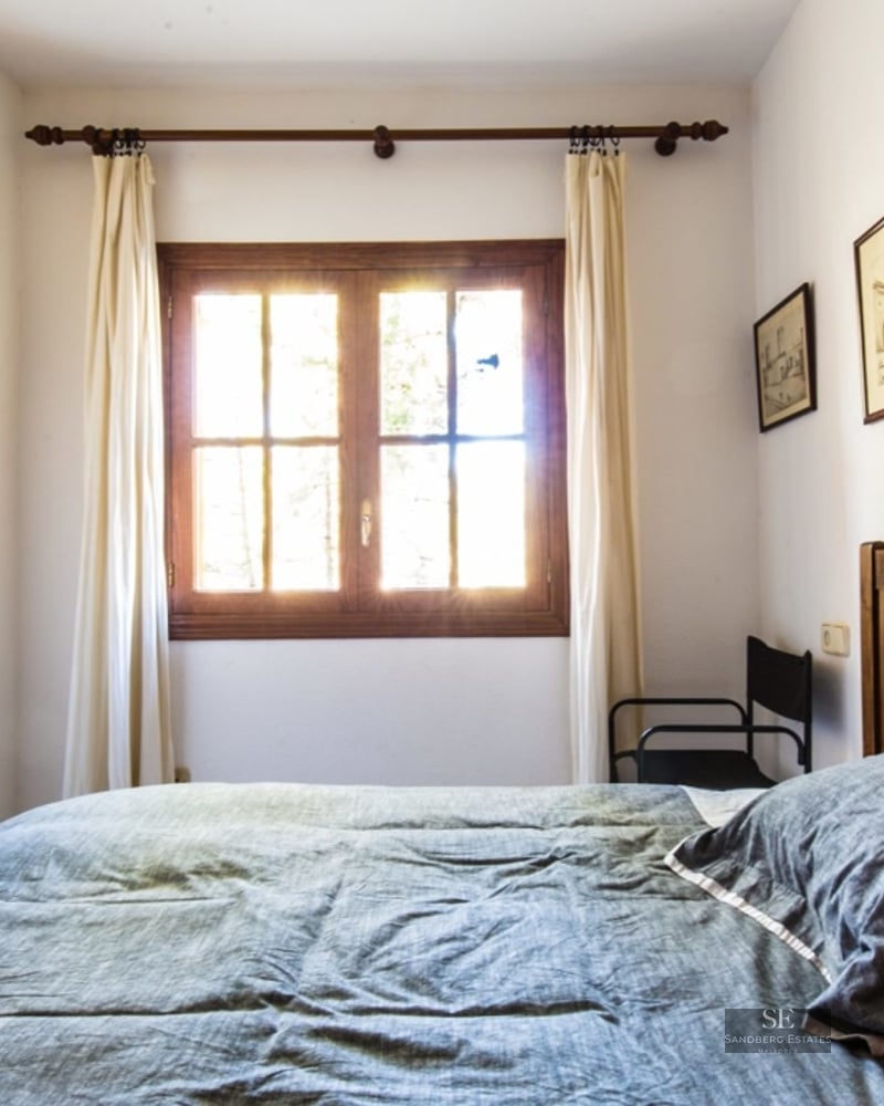 A bedroom with a wooden bed, grey bedding, and a window with bright sunlight coming through white curtains.