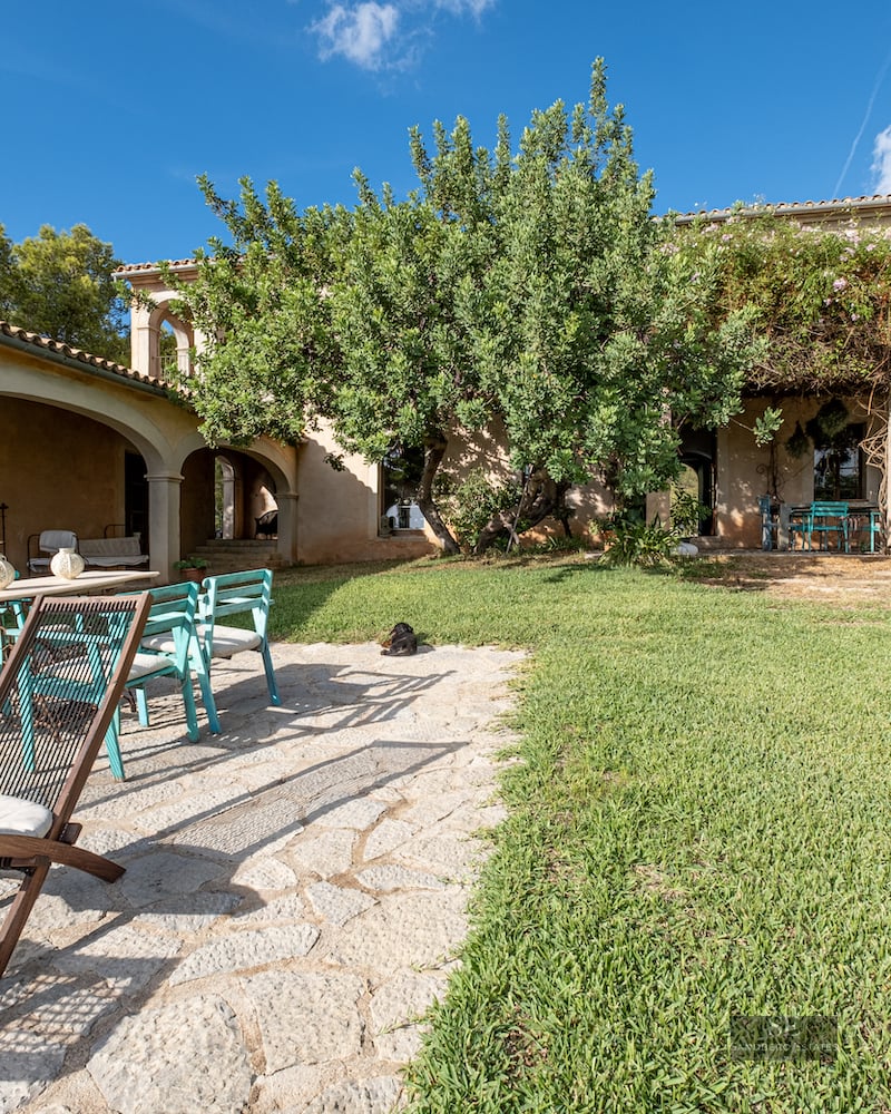 A sunny garden with a stone patio, lawn, teal chairs, wooden loungers, and a rustic villa with arched porches.