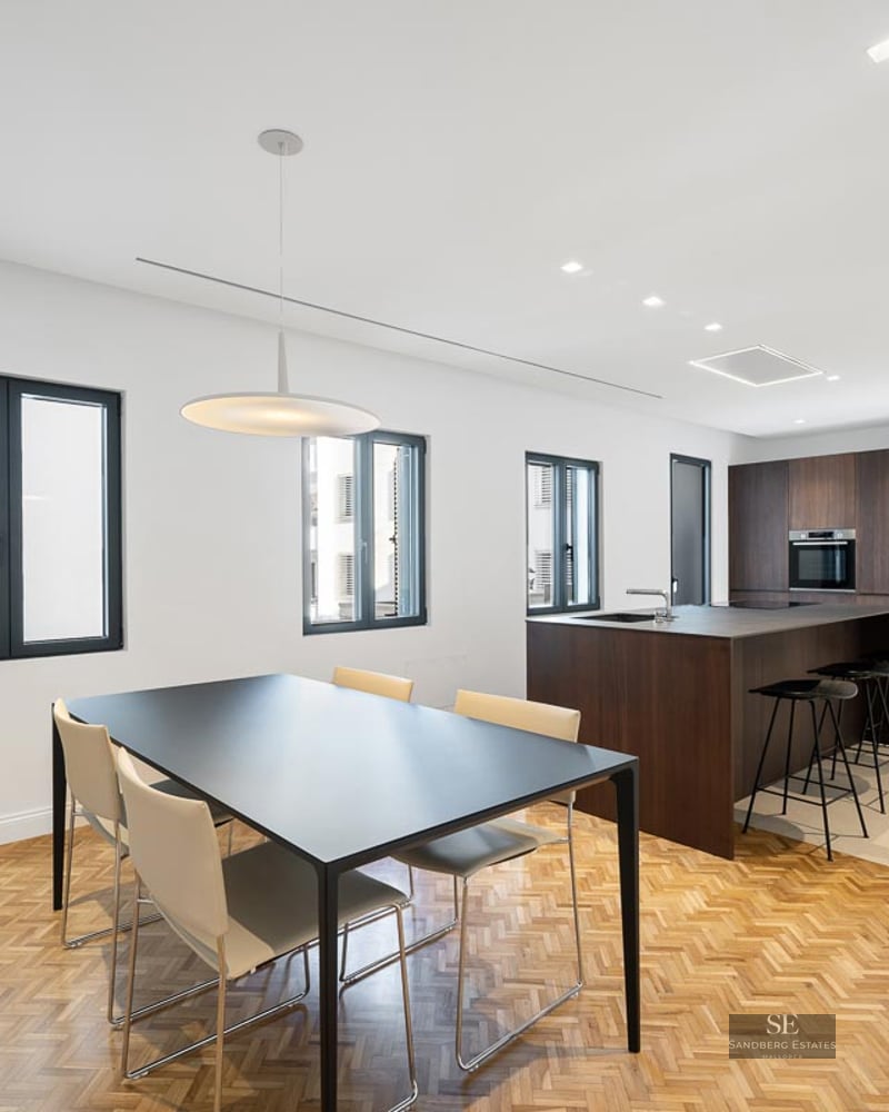 Modern kitchen with dark wood island and dining area featuring herringbone wood floors and large windows.
