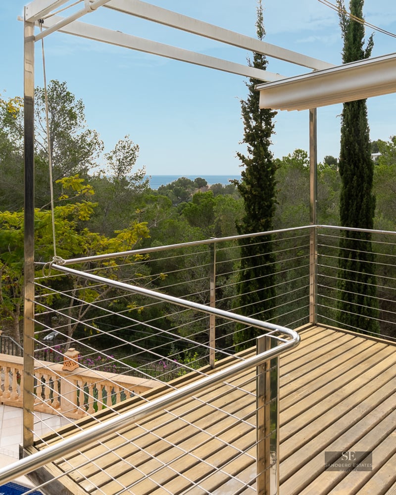 Elevated wooden deck with metal railings overlooking a pool and lush green forest under a blue sky.