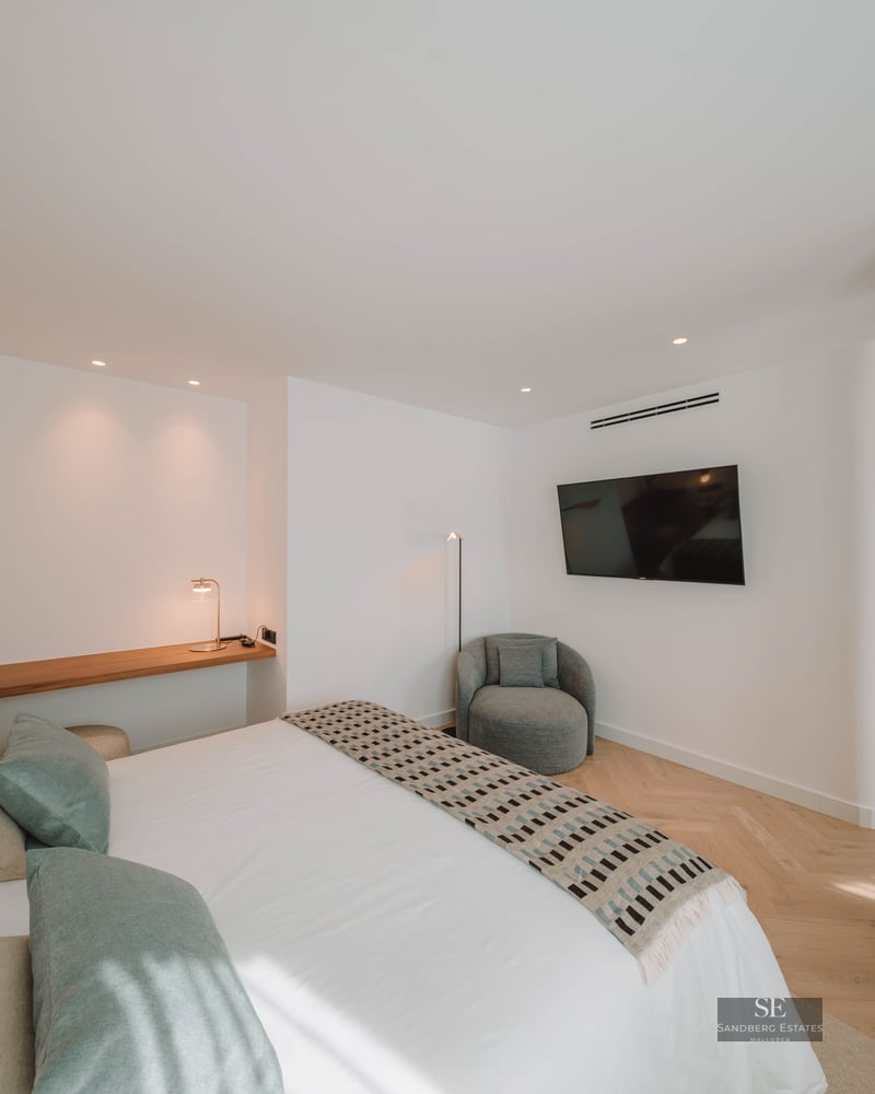 Modern bedroom featuring a white bed with sage green pillows, wooden shelf, and herringbone wood flooring.
