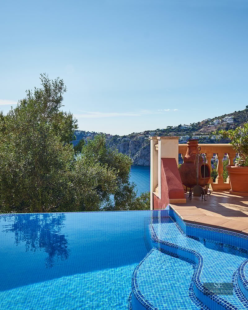 An infinity pool with blue mosaic tiles overlooking a coastal bay and mountains under a clear blue sky.