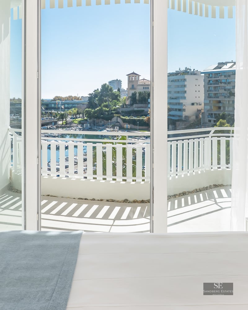 Bright bedroom with white bed, blue pillow, and balcony overlooking a marina under a clear blue sky.