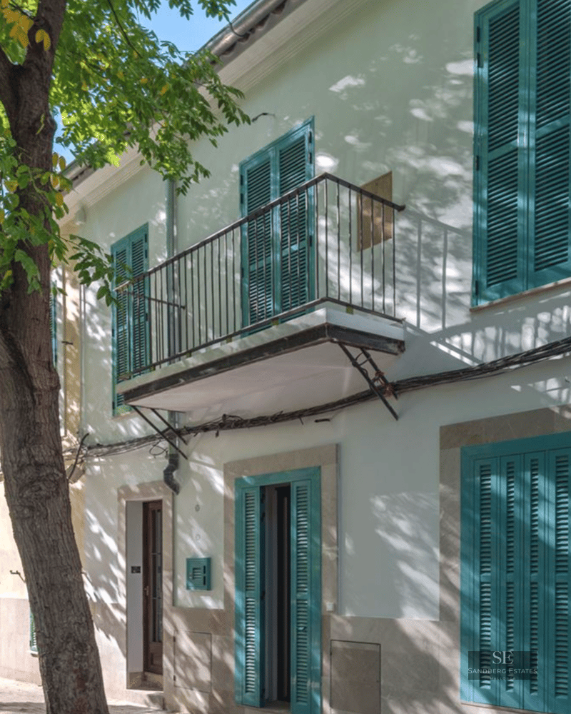 White building exterior featuring turquoise shutters, a small iron balcony, and shadows from a large leafy tree.
