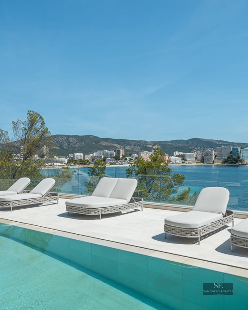 Modern white terrace with a turquoise pool, loungers, and a glass railing overlooking a scenic Mediterranean bay.