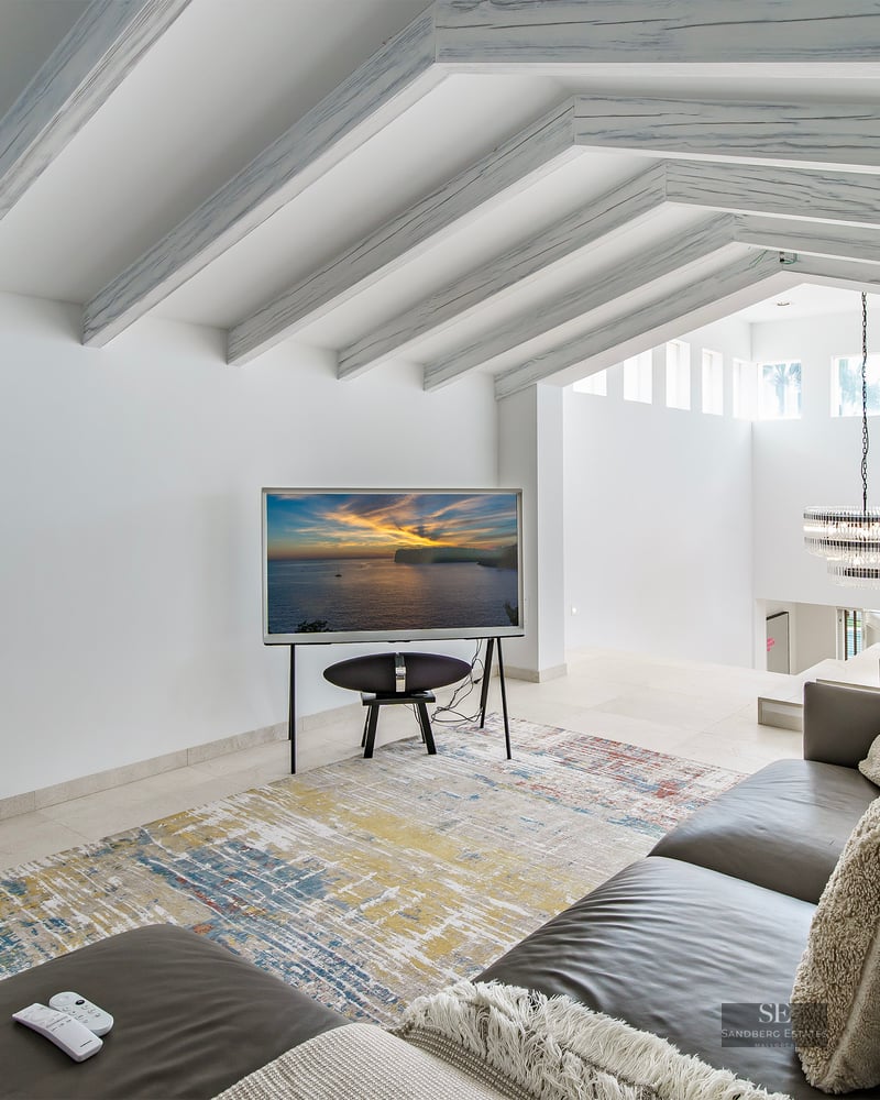 Bright living room featuring a grey leather sofa, abstract rug, and sloped ceiling with white-washed wooden beams.