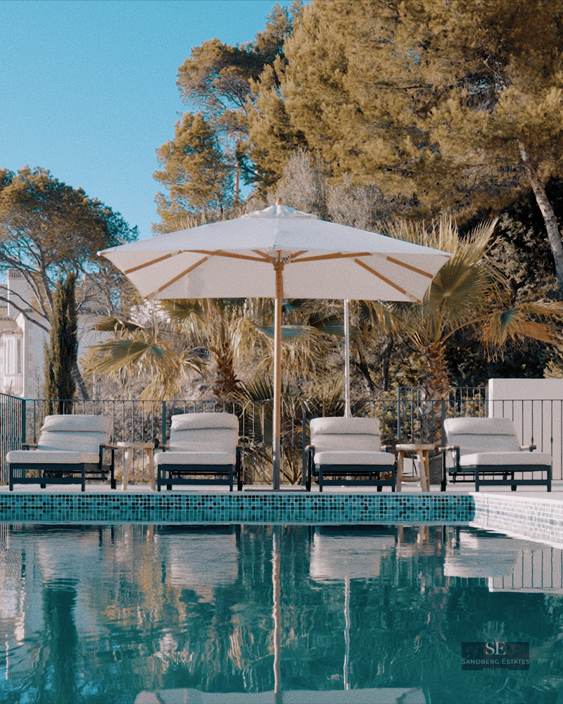 Infinity swimming pool with four sunbeds under a white umbrella, surrounded by stone walls and Mediterranean greenery.
