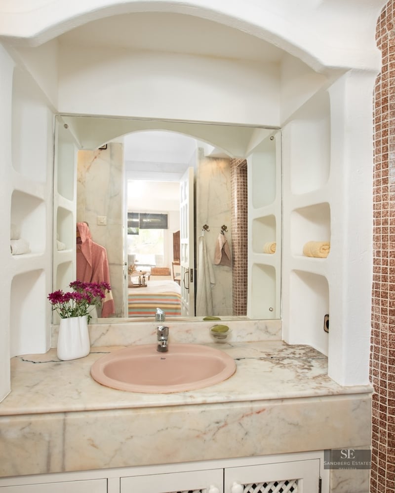 Bathroom vanity with pink sink, marble countertop, arched mirror, and brown mosaic tile pillar.