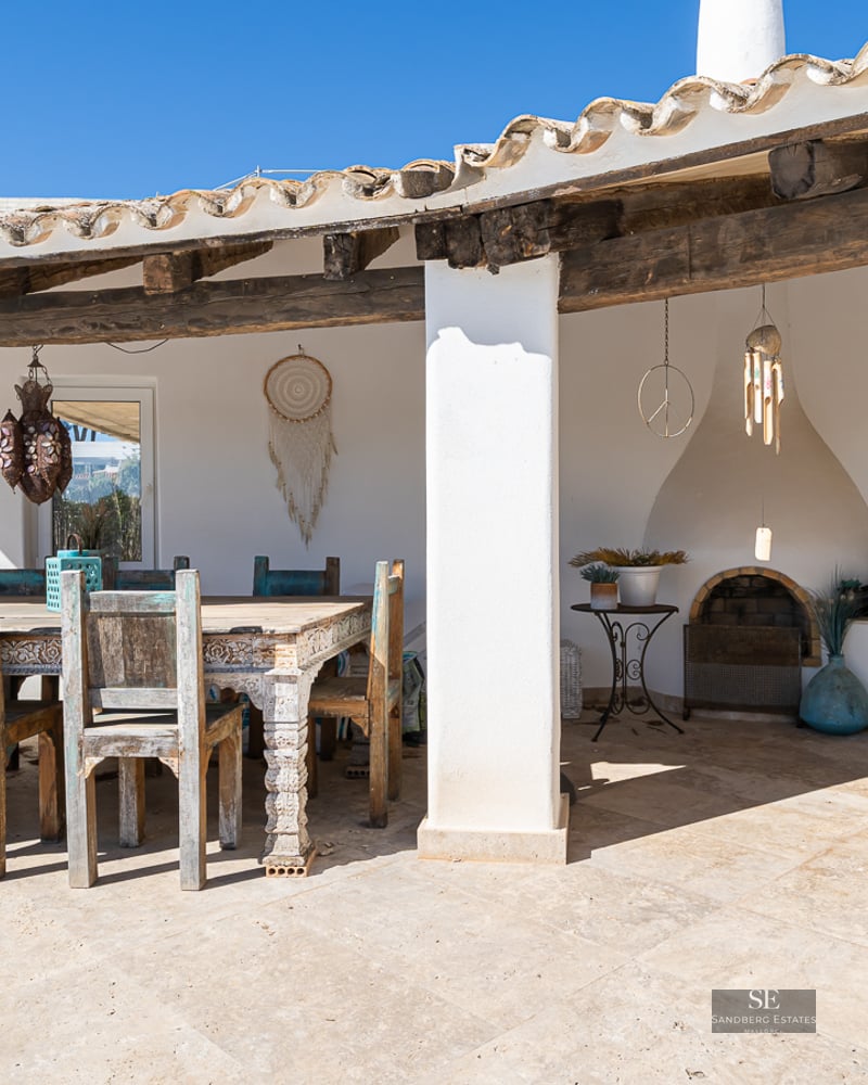 A covered terrace with a rustic wooden dining set, stone flooring, a built-in fireplace, and Mediterranean decor.