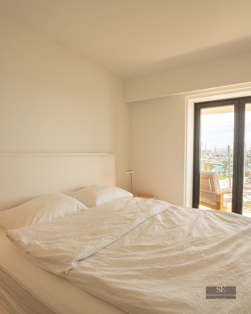 Bright minimalist bedroom with white linens, a grey chair, and large glass doors overlooking a marina with boats.