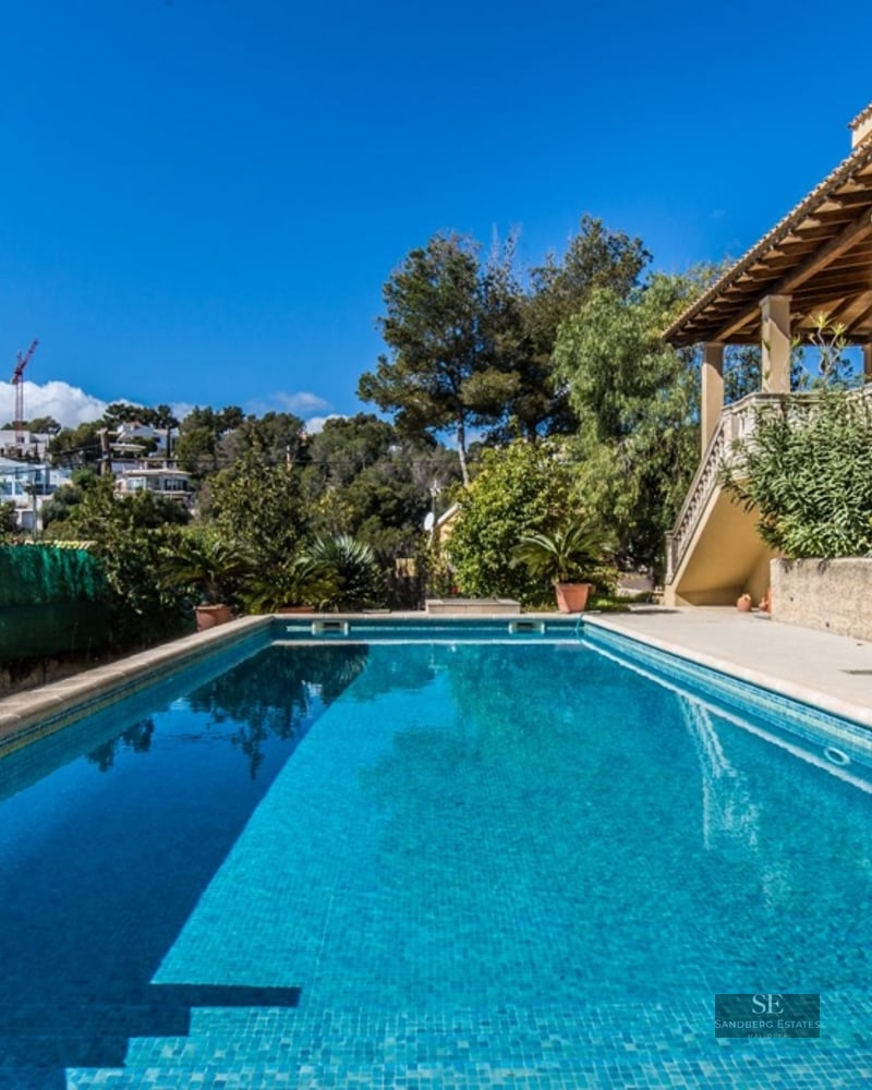 Large rectangular swimming pool with turquoise water next to a Mediterranean villa with a sunny terrace and blue sky.