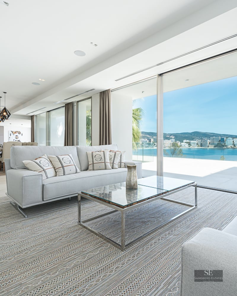 Modern living room with grey sofas and large glass doors opening to a panoramic view of the sea and coastline.