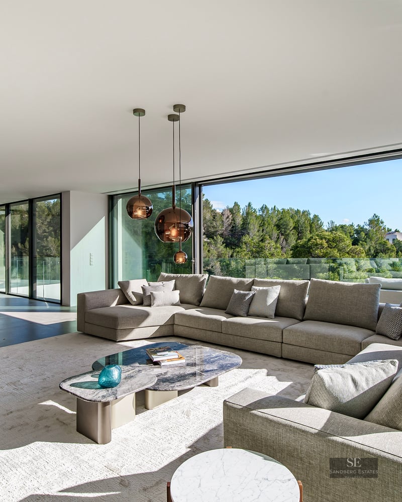 Large modern living room with floor-to-ceiling windows, a grey L-shaped sofa, and views of a green forest.