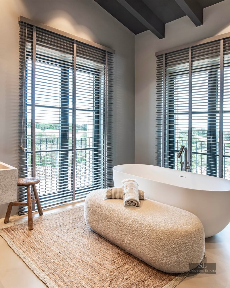 Modern master bathroom featuring a freestanding white tub, large stone sink, and windows with grey blinds.