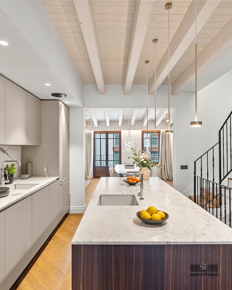 Contemporary kitchen featuring a large marble island, beige cabinetry, wooden ceiling beams, and a black staircase.