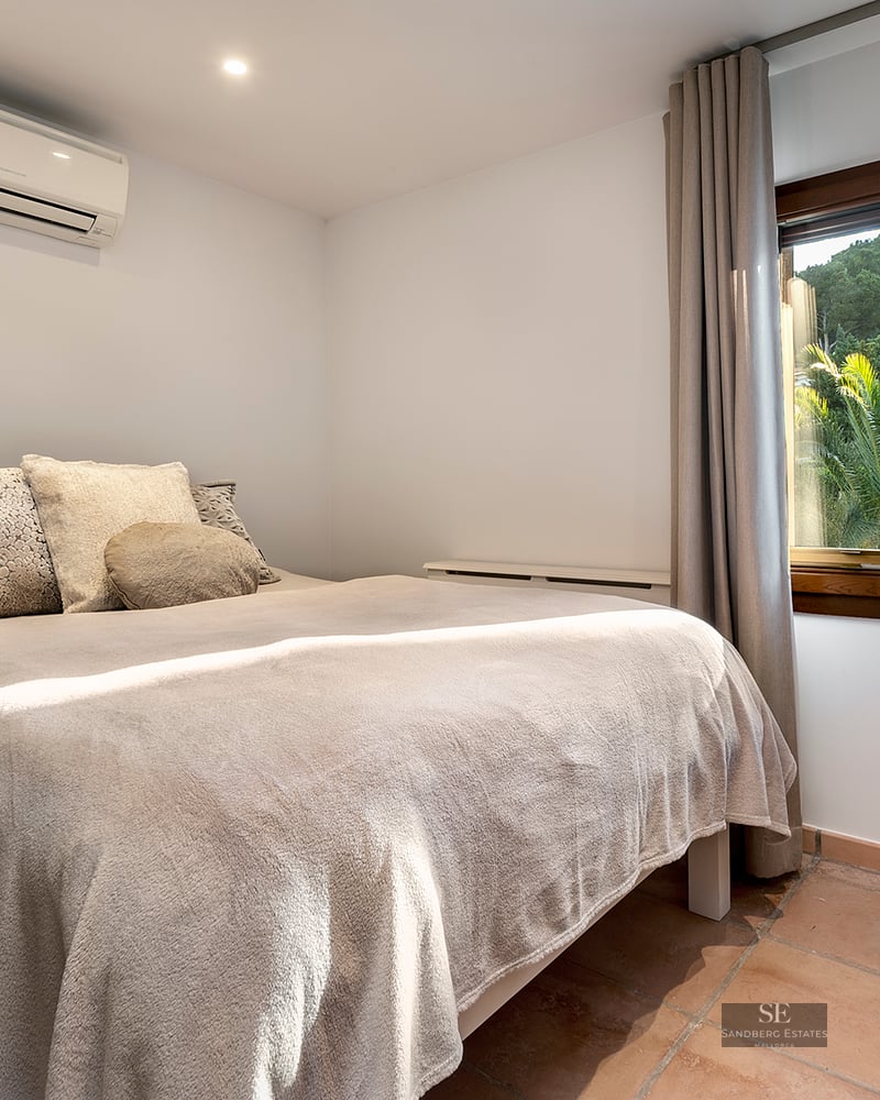 Bedroom with a cozy double bed, terracotta floors, air conditioning, and a window overlooking lush green palm trees.