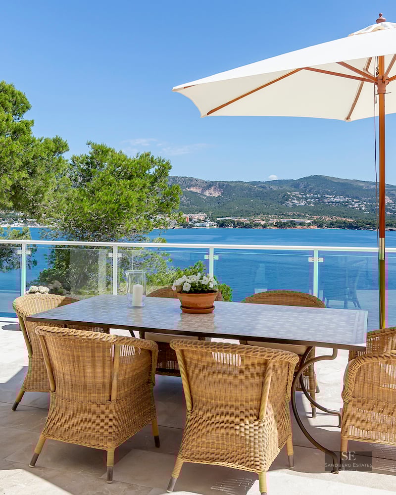 Outdoor dining area with wicker chairs and white umbrella on a stone terrace overlooking the blue sea and mountains.