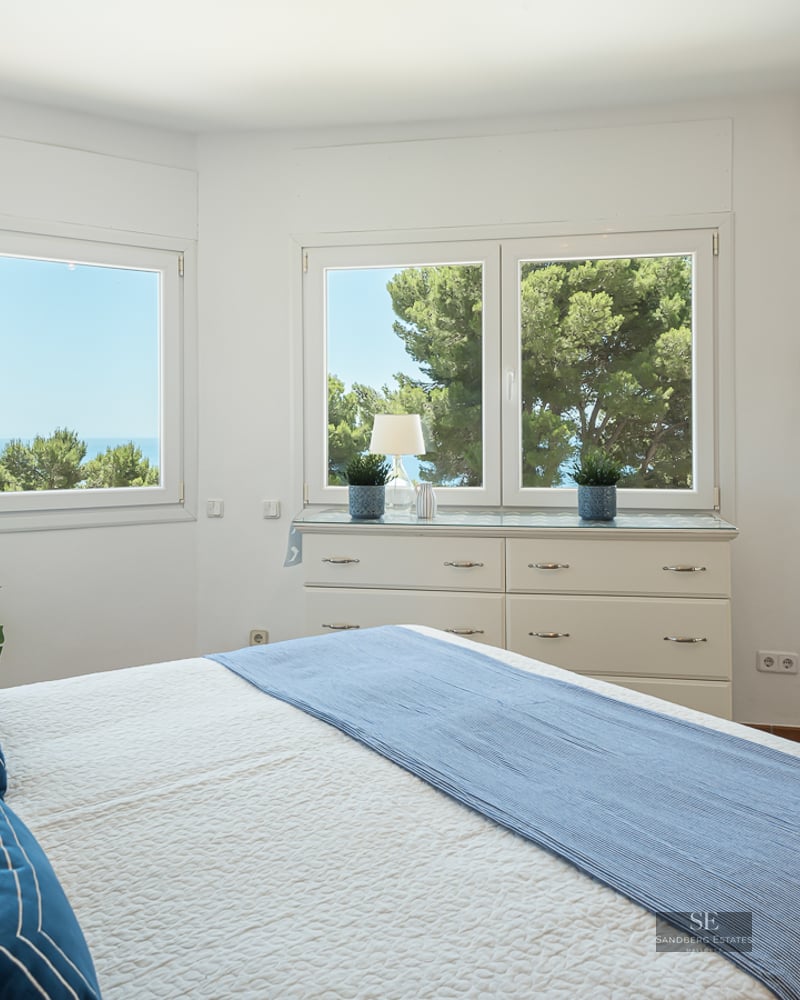 Bright white bedroom with large windows overlooking the sea, blue bedding, and a door to a sunny balcony.
