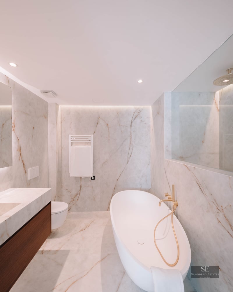 Modern marble bathroom featuring a white freestanding tub, gold fixtures, and an integrated double vanity.