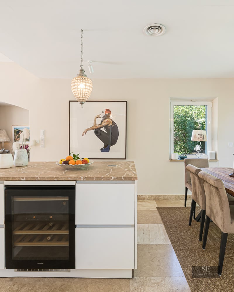 A modern white kitchen island with a wine fridge next to a rustic wooden dining table overlooking a lush garden.