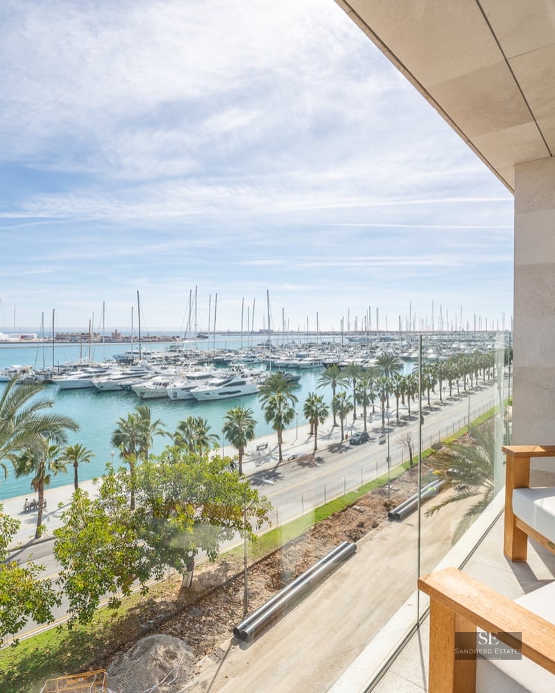 Modern balcony with wooden chairs and glass railing overlooking a marina full of yachts and palm trees.