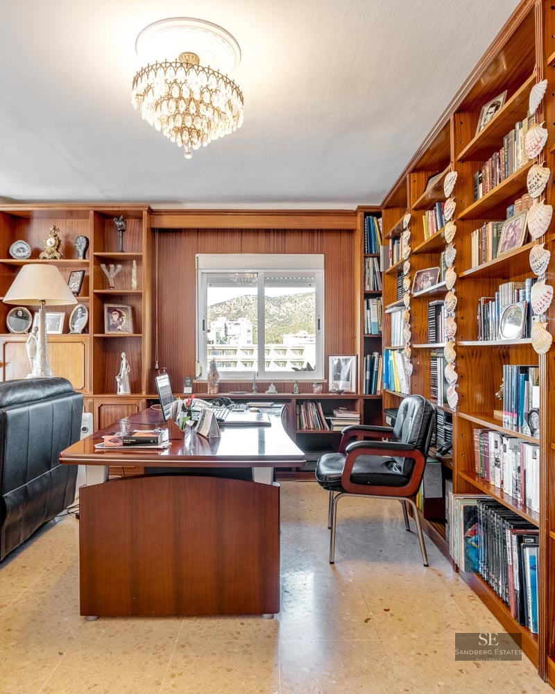 Spacious home office featuring floor-to-ceiling wooden bookshelves, a large desk, leather seating, and a crystal chandelier.