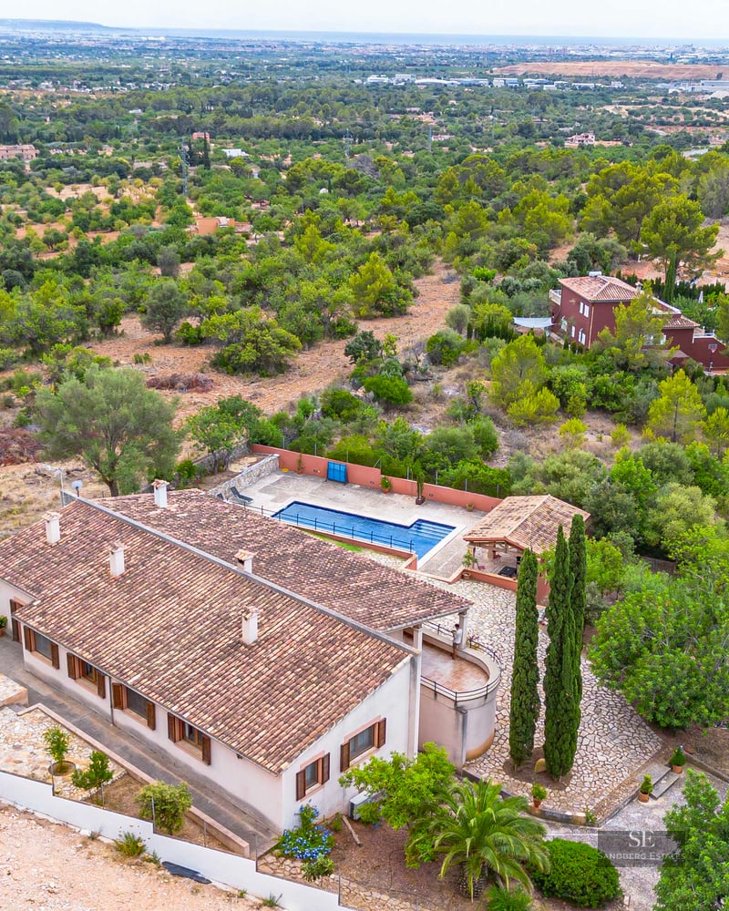 Bird's eye view of a large villa with a terracotta roof, a blue swimming pool, and lush green landscaped grounds.