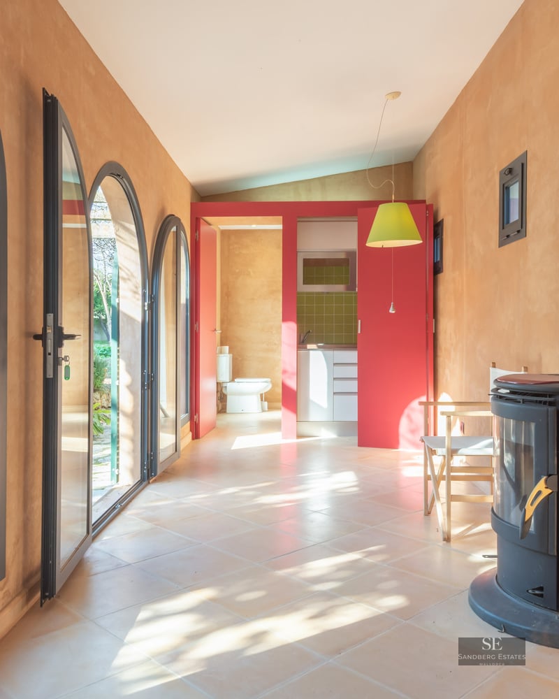 Sunlit room with terracotta walls, arched glass doors, a red wood-burning stove, and a view to an outdoor patio.