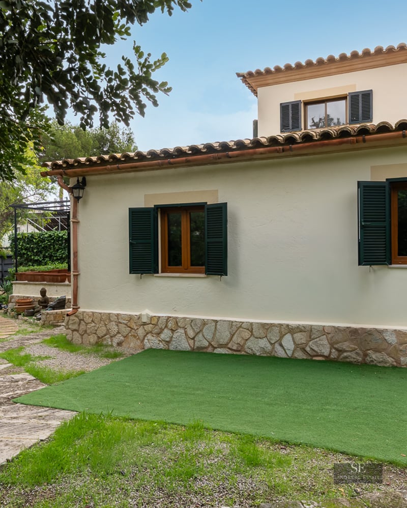 Cream stucco villa with green shutters, stone base, and a winding stone garden path under a clear blue sky.