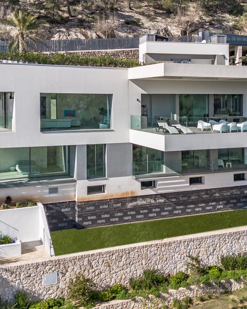 Modern white multi-story villa with large glass windows and terraces built into a rocky hillside with stone walls.