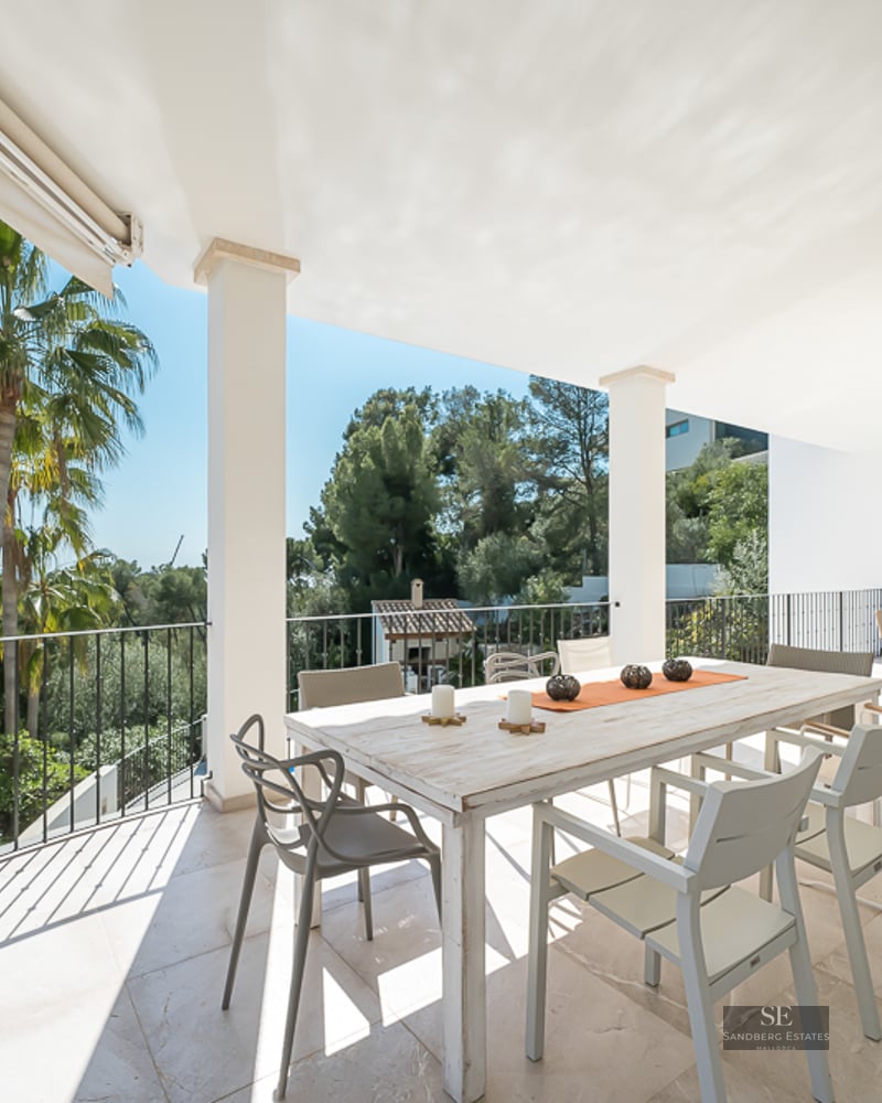 Terrasse blanche avec table à manger en bois, chaises modernes et vue sur les palmiers et la mer.