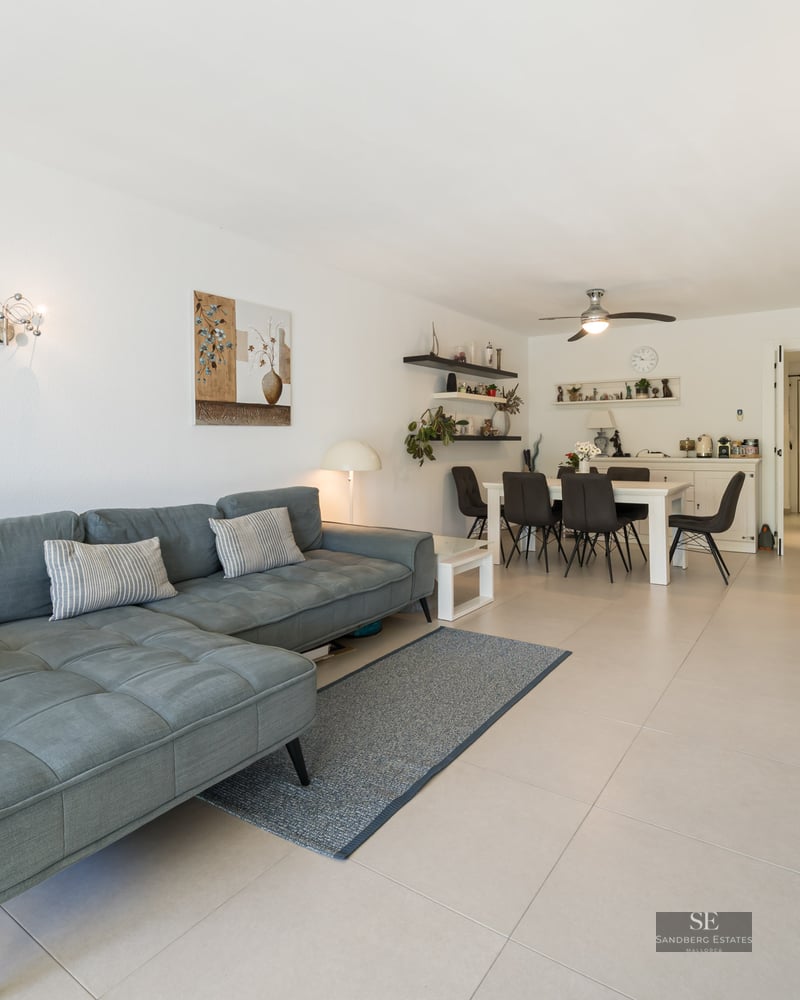 Salon lumineux avec un grand canapé en L bleu, des meubles blancs, un coin repas et un sol carrelé.