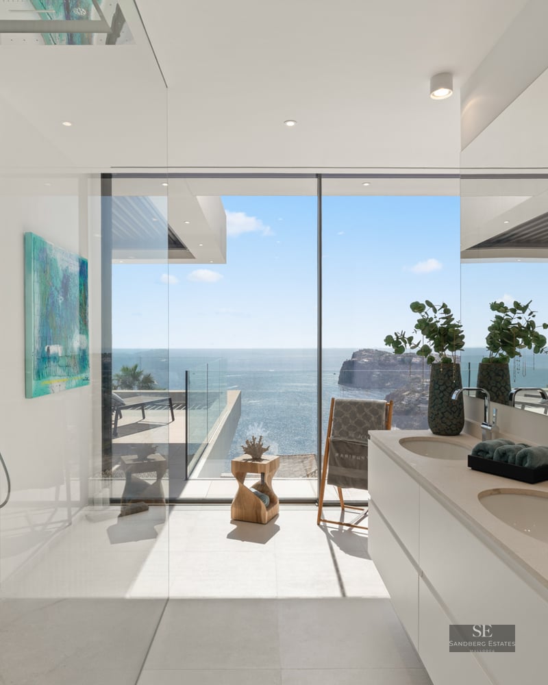Modern white bathroom with glass walk-in shower and double vanity overlooking the ocean through floor-to-ceiling windows.