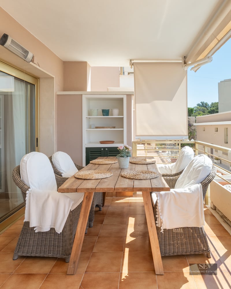 A sunlit balcony terrace with a wooden dining table, wicker chairs, and a metal railing overlooking urban buildings.