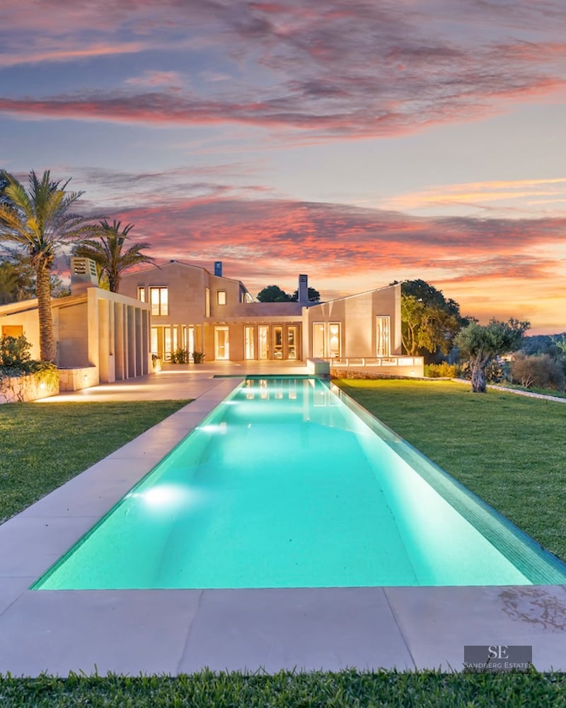 A long, illuminated rectangular pool leading to a modern stone villa under a vibrant pink and orange sunset sky.