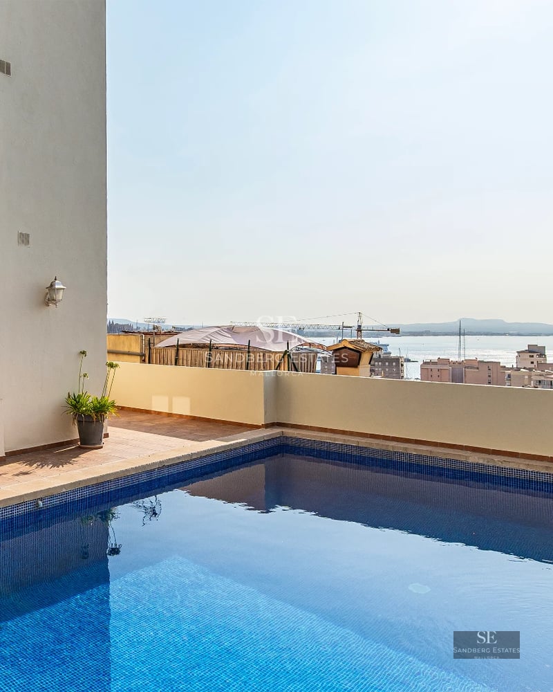 View of infinity pool with curved edge. Green garden, modern white house with large windows. Blue sky reflected in water.