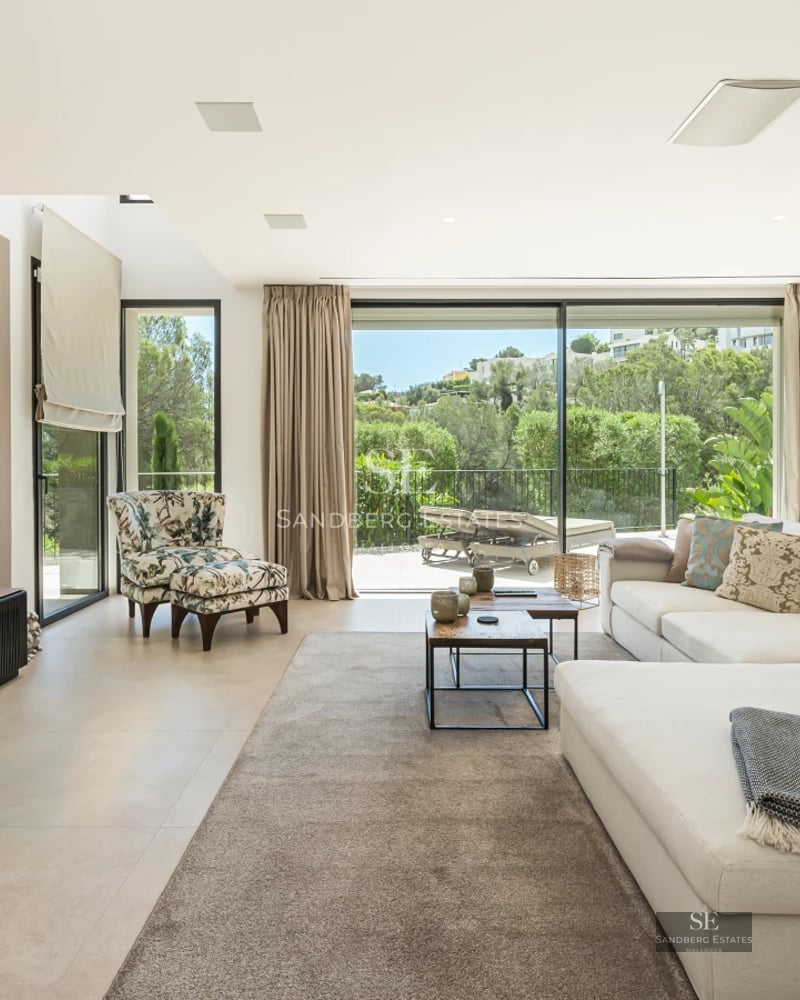 Spacious living room with floor-to-ceiling windows, light wood floors and minimalist design furniture. Views of pool and lush garden.