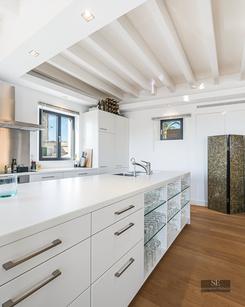 Modern white kitchen featuring a large island, wood flooring, ceiling beams, and a decorative wood sculpture.