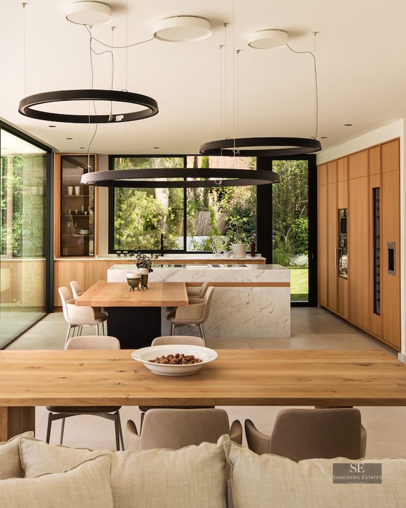High-end kitchen with a white marble island, wooden cabinets, and a long dining table under circular halo pendant lights.