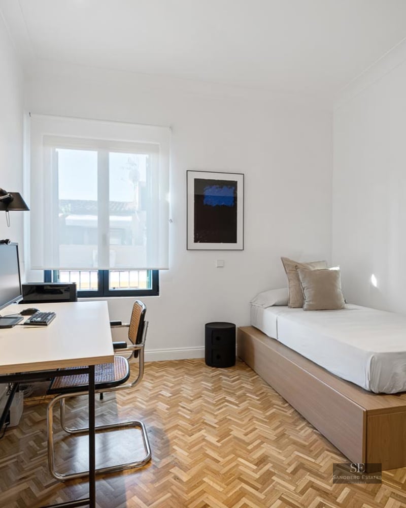 Bright minimalist room featuring a single bed, herringbone parquet floors, and a desk with a computer.