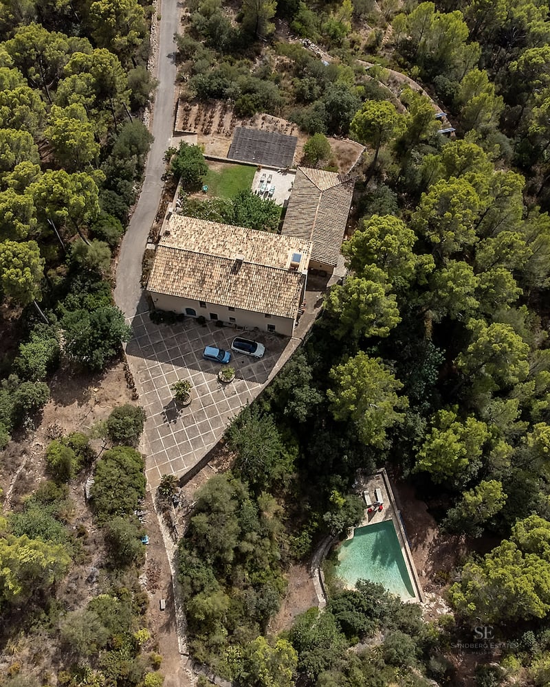 Aerial view of a stone house with terracotta roof, courtyard, and a pool, all surrounded by a dense green pine forest.