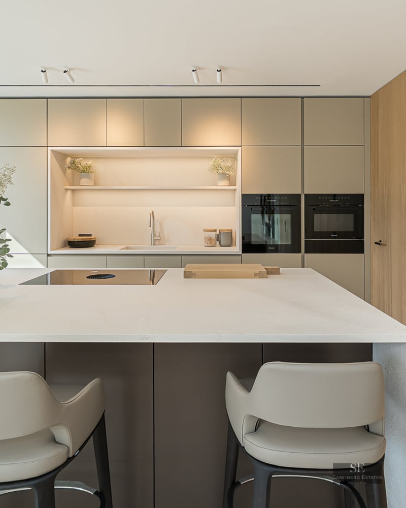 Modern kitchen island with stools, built-in appliances, wooden wall, and large window overlooking the water.